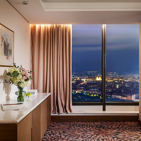 Grand Hotel Millennium 索菲亞 外观 照片 The photo depicts a modern hotel room or a lounge area with a stunning view of a cityscape at night. On the left, there's a wooden console table with a vase of flowers and a bottle of water. A piece of art is framed on the wall above the table. The r
