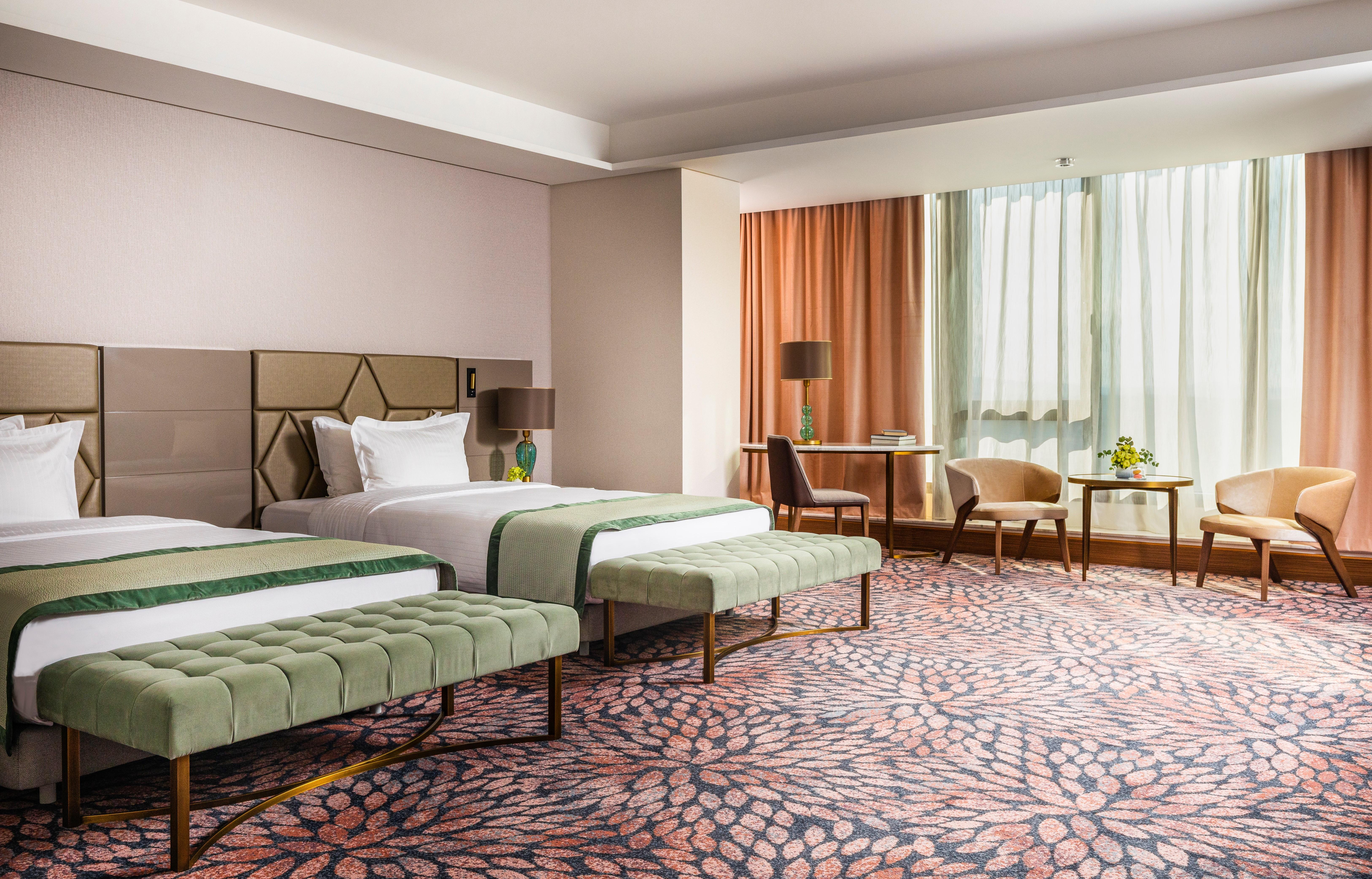 Grand Hotel Millennium 索菲亞 外观 照片 The image shows a well-decorated hotel room featuring two beds with green striped bedspreads. There is a plush green bench at the foot of each bed. The room has large windows with sheer curtains allowing natural light to enter. There is a cozy seatin