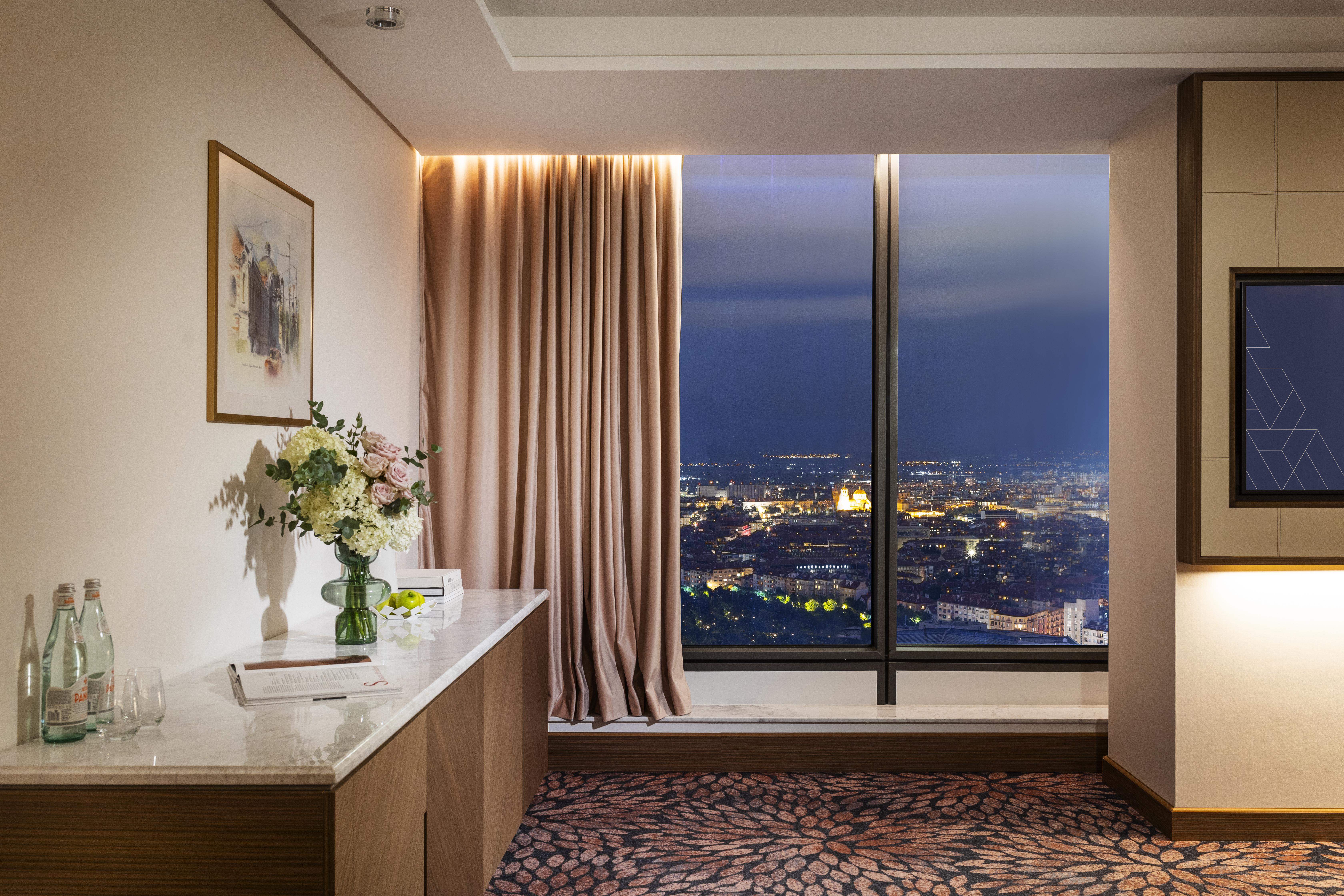 Grand Hotel Millennium 索菲亞 外观 照片 The photo depicts a modern hotel room or a lounge area with a stunning view of a cityscape at night. On the left, there's a wooden console table with a vase of flowers and a bottle of water. A piece of art is framed on the wall above the table. The r