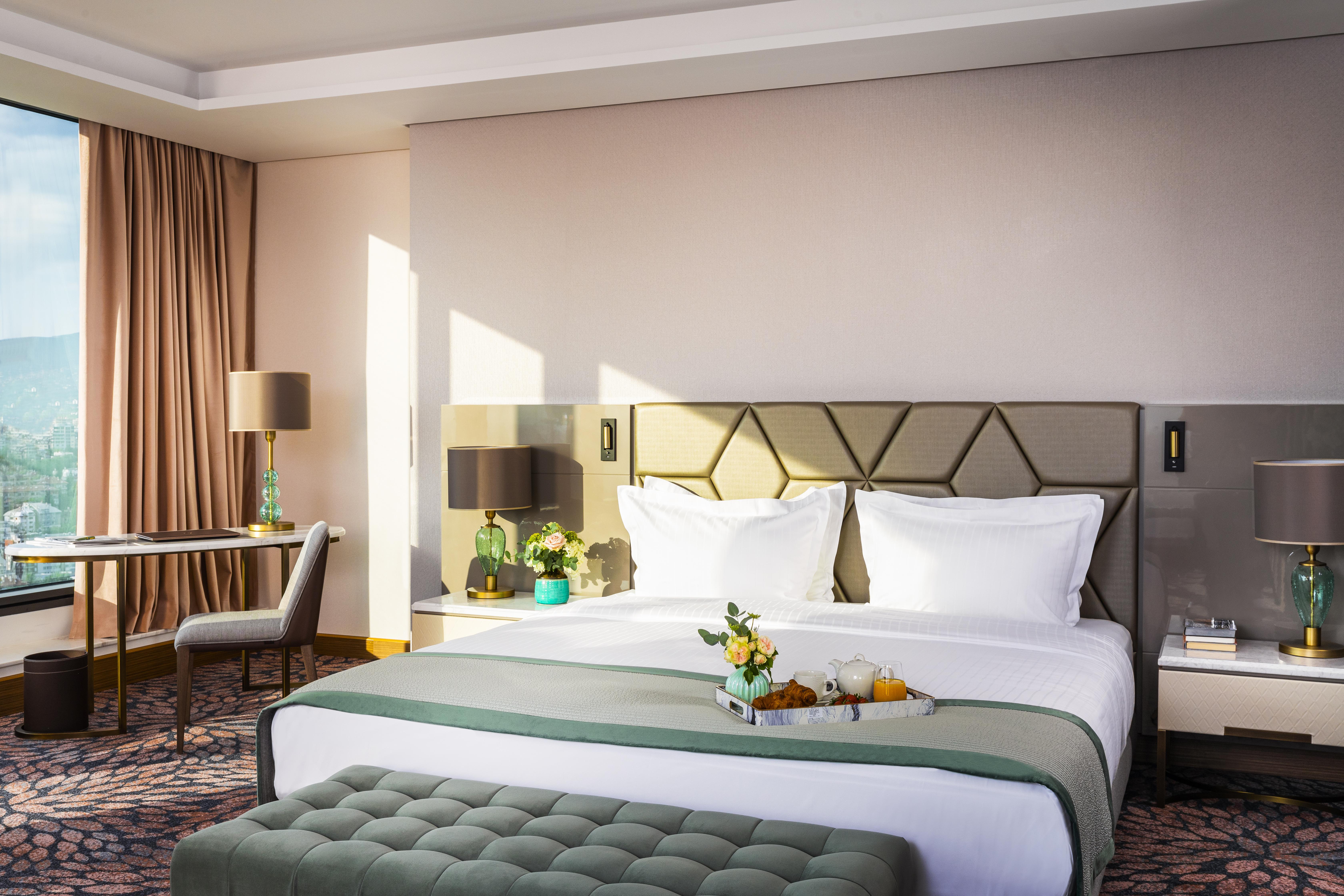 Grand Hotel Millennium 索菲亞 外观 照片 The photo depicts a stylish hotel room featuring a large bed with white linens and a green decorative throw. Behind the bed is a geometric-patterned headboard. To the side, there are modern bedside lamps and a small table with a vase of flowers. A se
