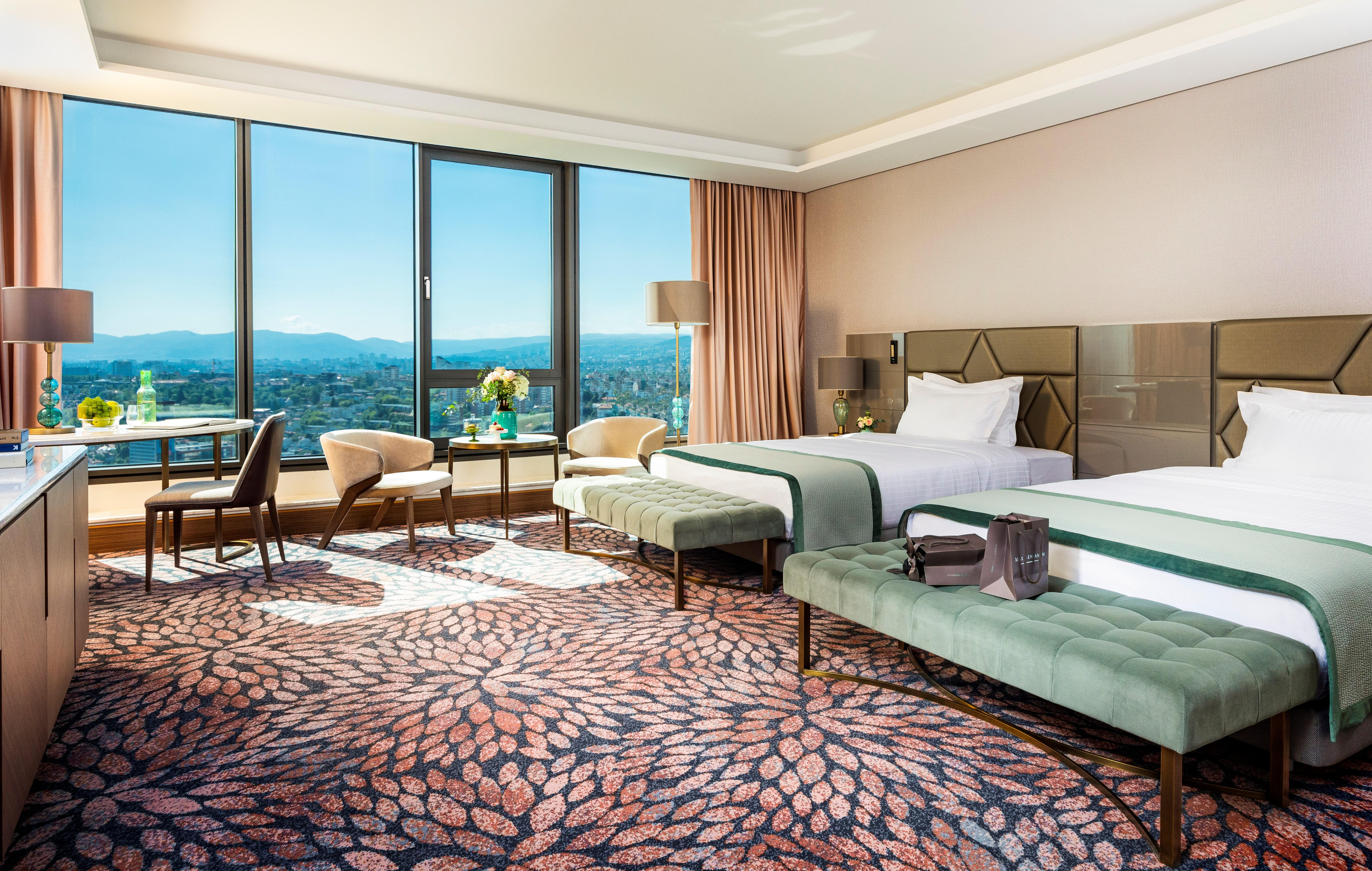 Grand Hotel Millennium 索菲亞 外观 照片 The photo depicts a luxurious hotel room featuring two beds with elegant bedding. The room has large windows that offer a panoramic view of the city and mountains in the background. There is a decorative area rug with a floral pattern covering the fl
