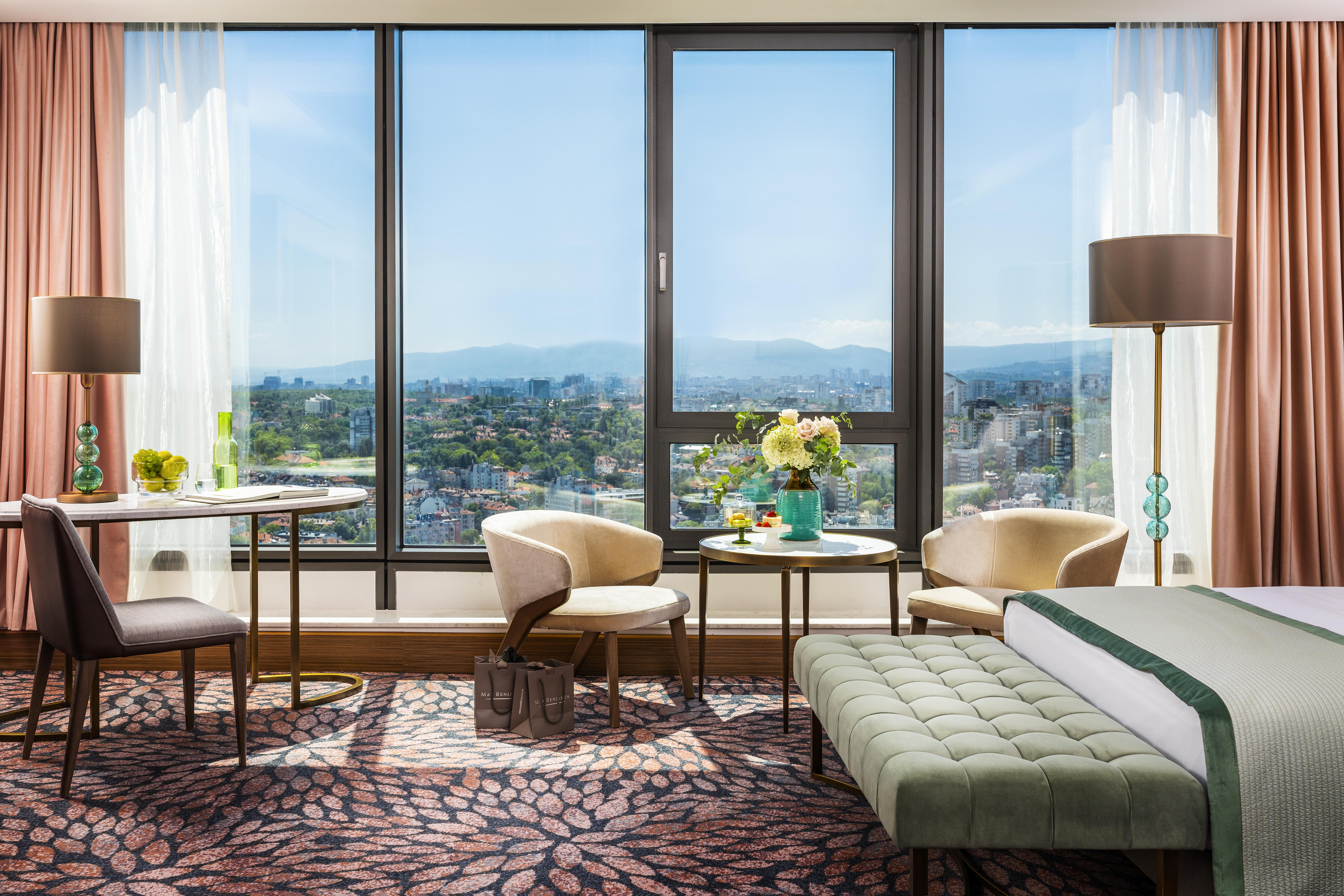 Grand Hotel Millennium 索菲亞 外观 照片 The photo shows a modern and well-designed hotel room featuring a large window that offers a view of the outdoors, including mountains in the background. Inside the room, there is a neatly made bed with a green accent blanket, a stylish rug with a fl