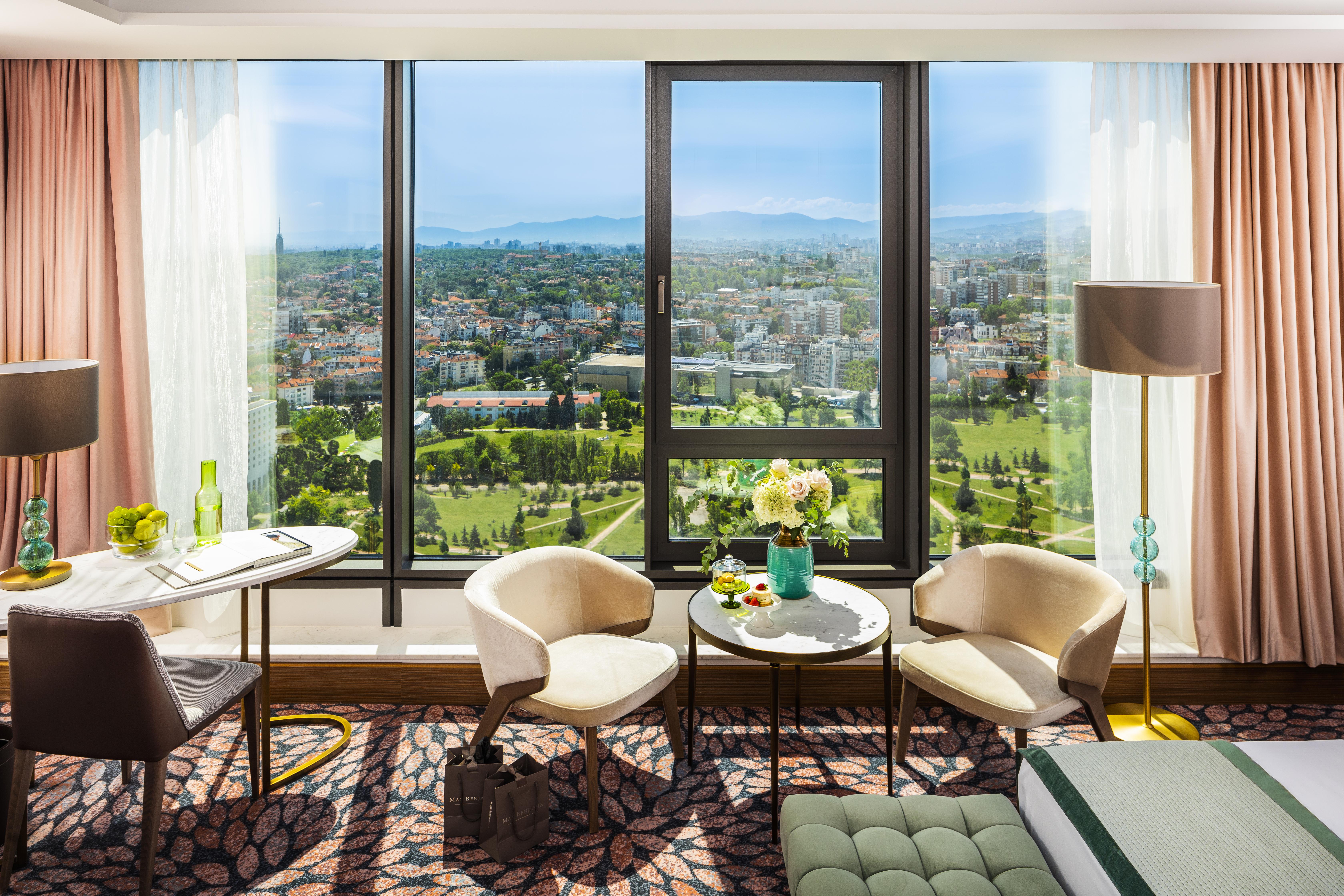 Grand Hotel Millennium 索菲亞 外观 照片 The photo shows a stylish hotel room with large windows providing a beautiful view of a cityscape and landscape beyond. There are two modern chairs and a small round table set near the windows, accompanied by a vase of fresh flowers. The room feature