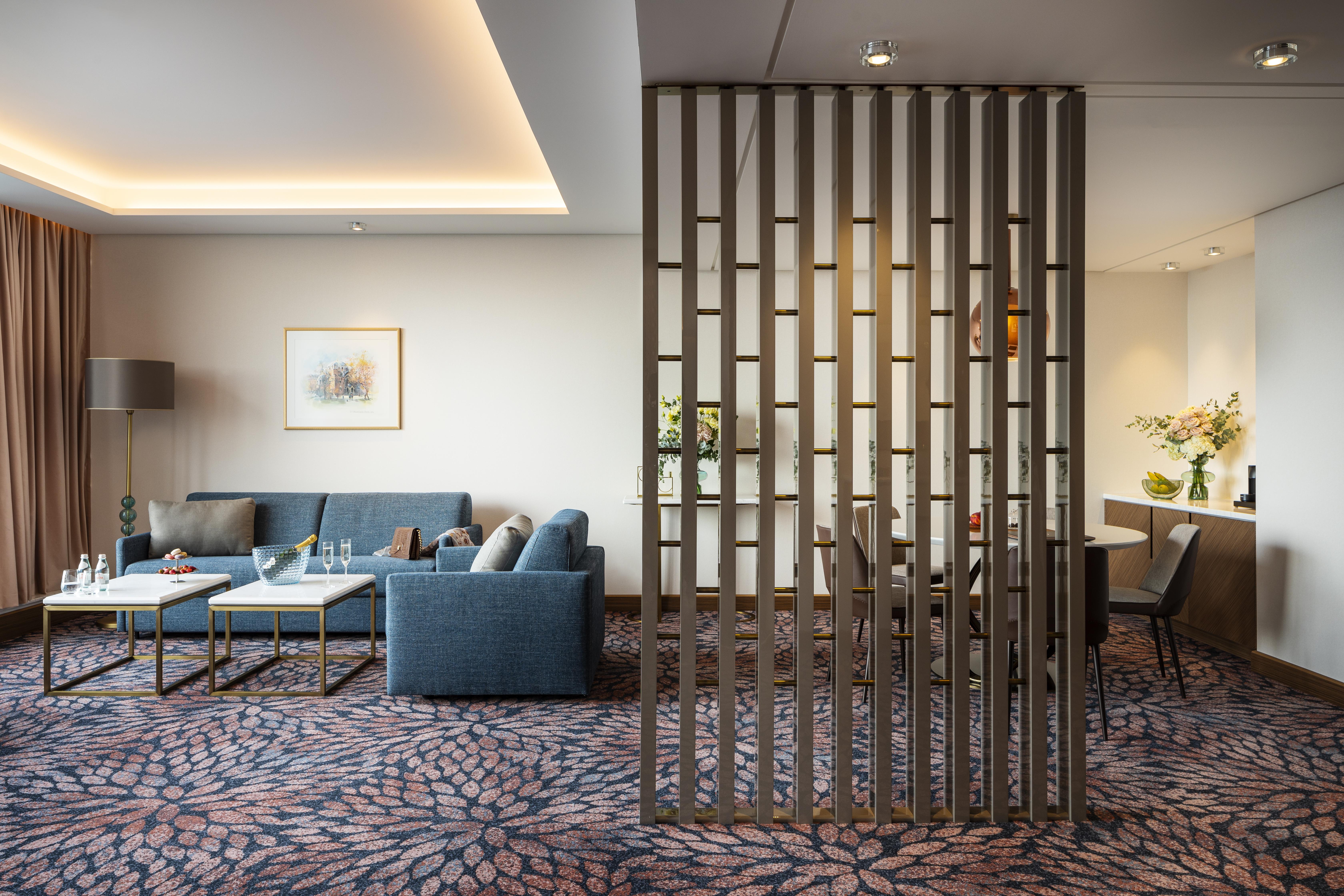 Grand Hotel Millennium 索菲亞 外观 照片 The photo shows a modern living space with a comfortable seating area featuring a blue couch and two coffee tables. There is a decorative partition made of vertical wooden slats, which separates this sitting area from a dining space. The dining area 