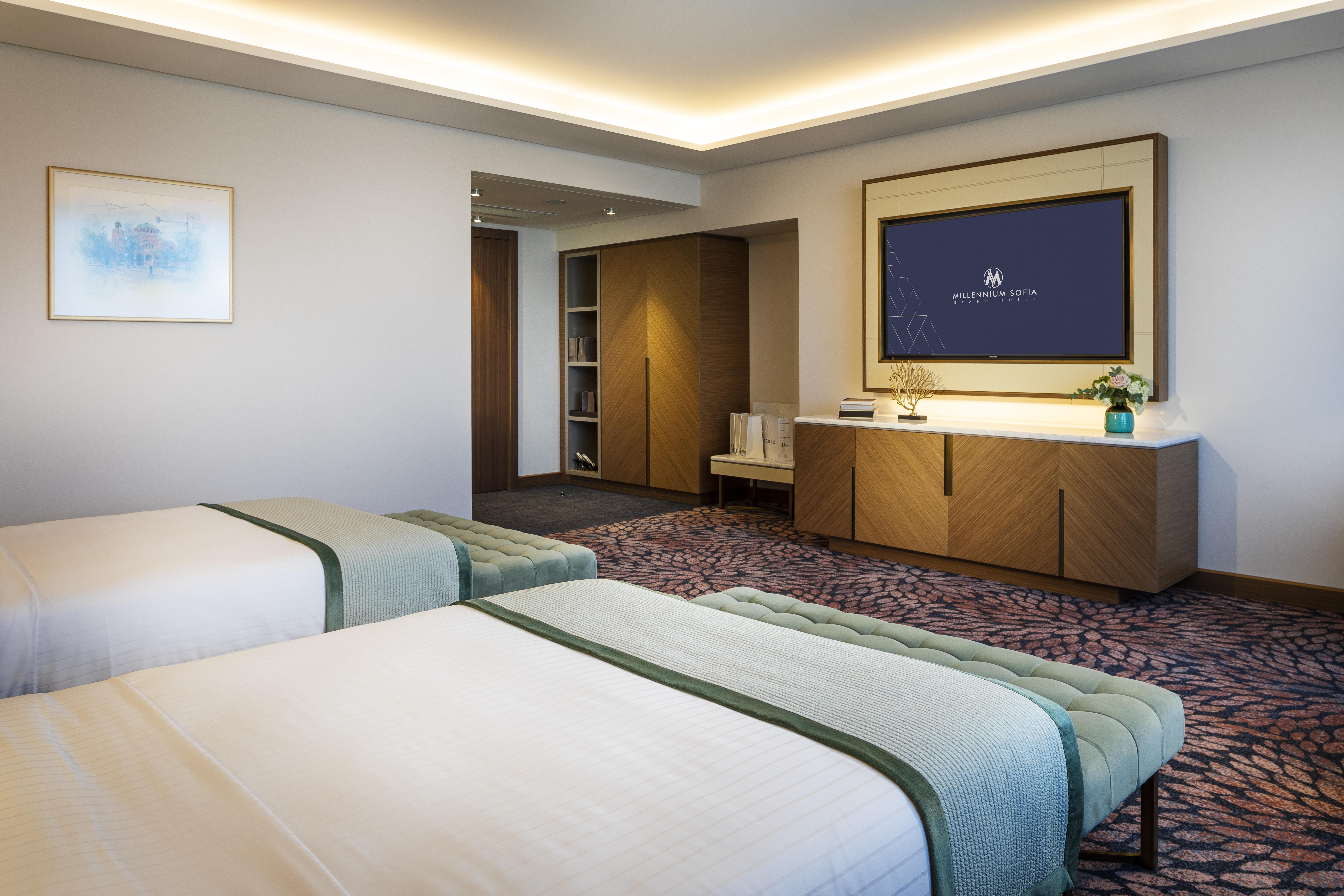Grand Hotel Millennium 索菲亞 外观 照片 The photo shows a modern hotel room featuring two beds with light-colored bedding and decorative throws. The room has a contemporary design, characterized by neutral walls and a plush carpet. A large television is mounted on the wall across from the 