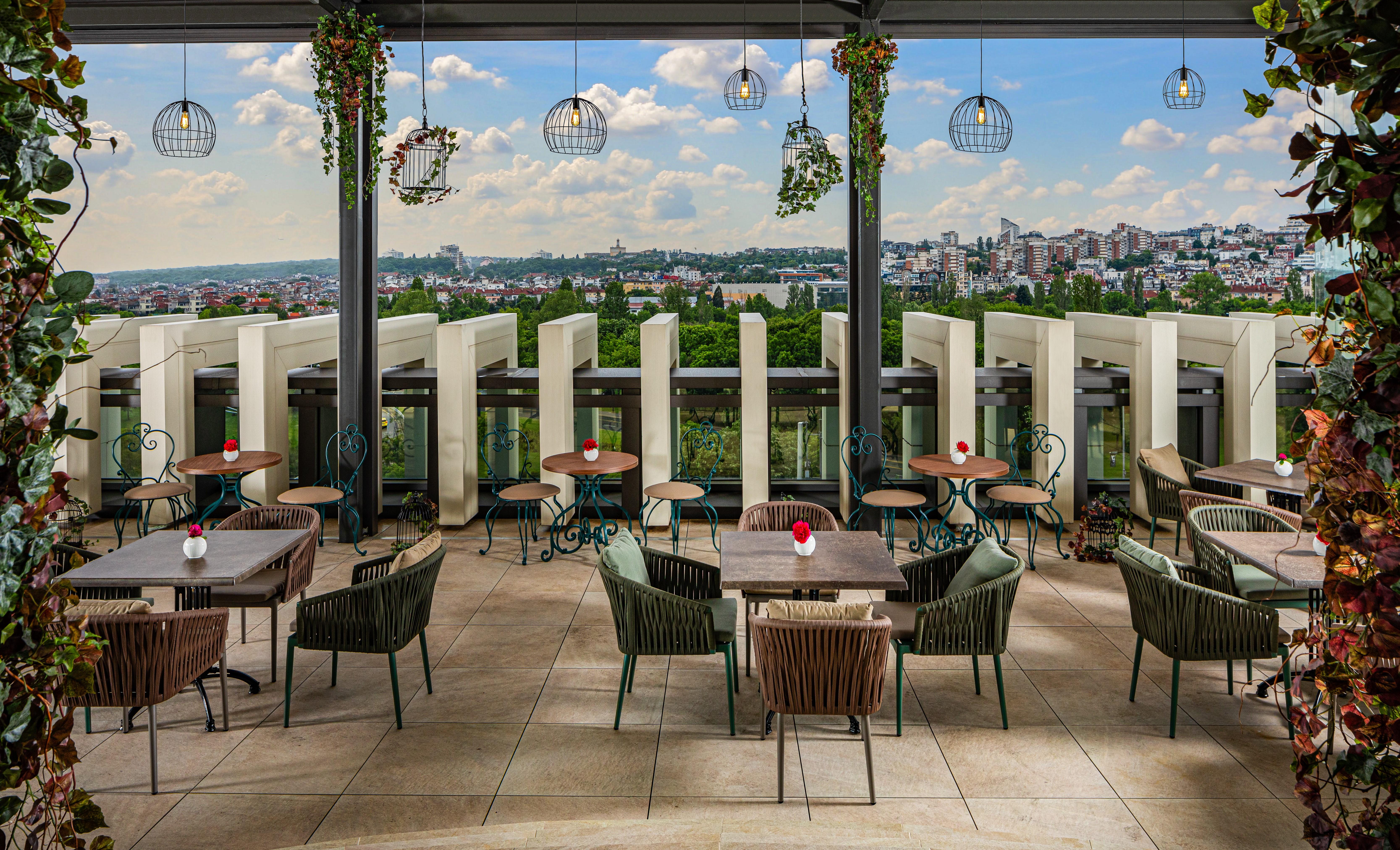 Grand Hotel Millennium 索菲亞 外观 照片 The photo shows a stylish outdoor restaurant or café setting. There are several tables arranged on a tiled patio, each with chairs, some made of woven materials. Centerpieces with small red flowers adorn each table. The space is partially shaded by o
