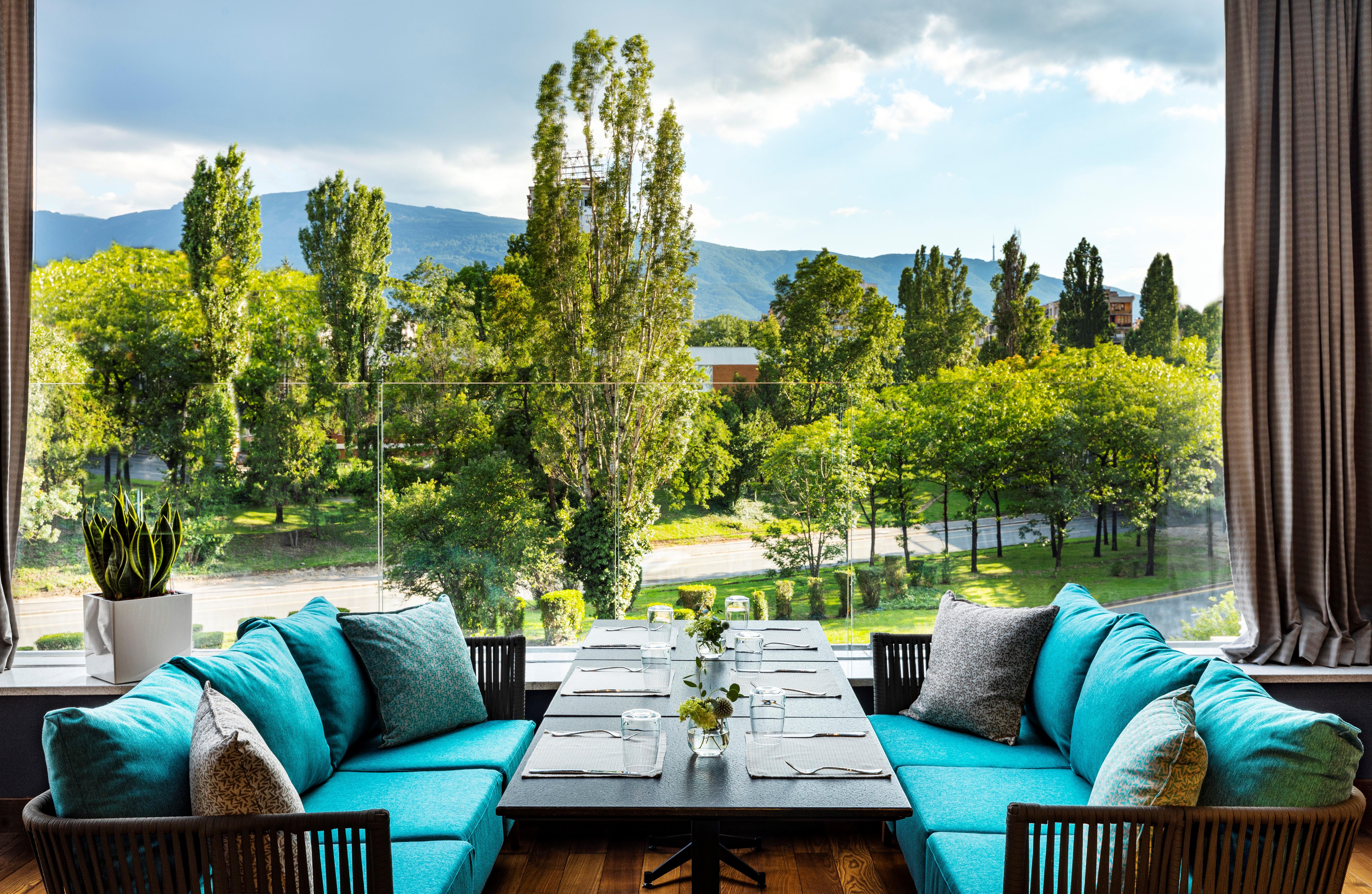 Grand Hotel Millennium 索菲亞 外观 照片 The photo shows a bright and inviting dining area with large windows that overlook a lush green landscape. There are several trees in the background, along with a view of distant mountains under a partly cloudy sky. The seating consists of stylish tu