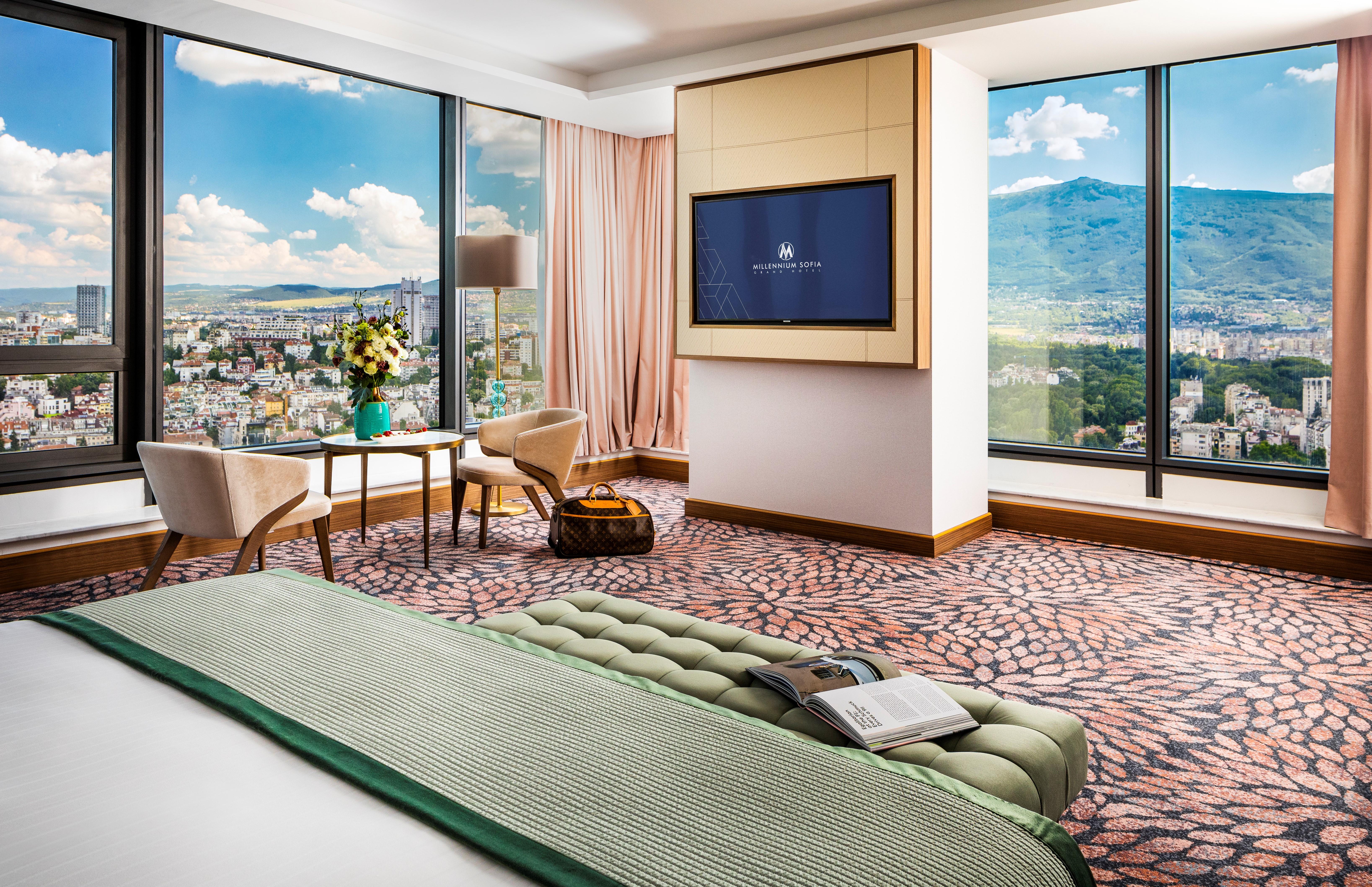 Grand Hotel Millennium 索菲亞 外观 照片 The photo showcases a stylish hotel room with modern decor. It features large windows providing a panoramic view of a cityscape and mountains in the background. Inside, there is a comfortable bed with a green bedspread, a seating area with two armcha