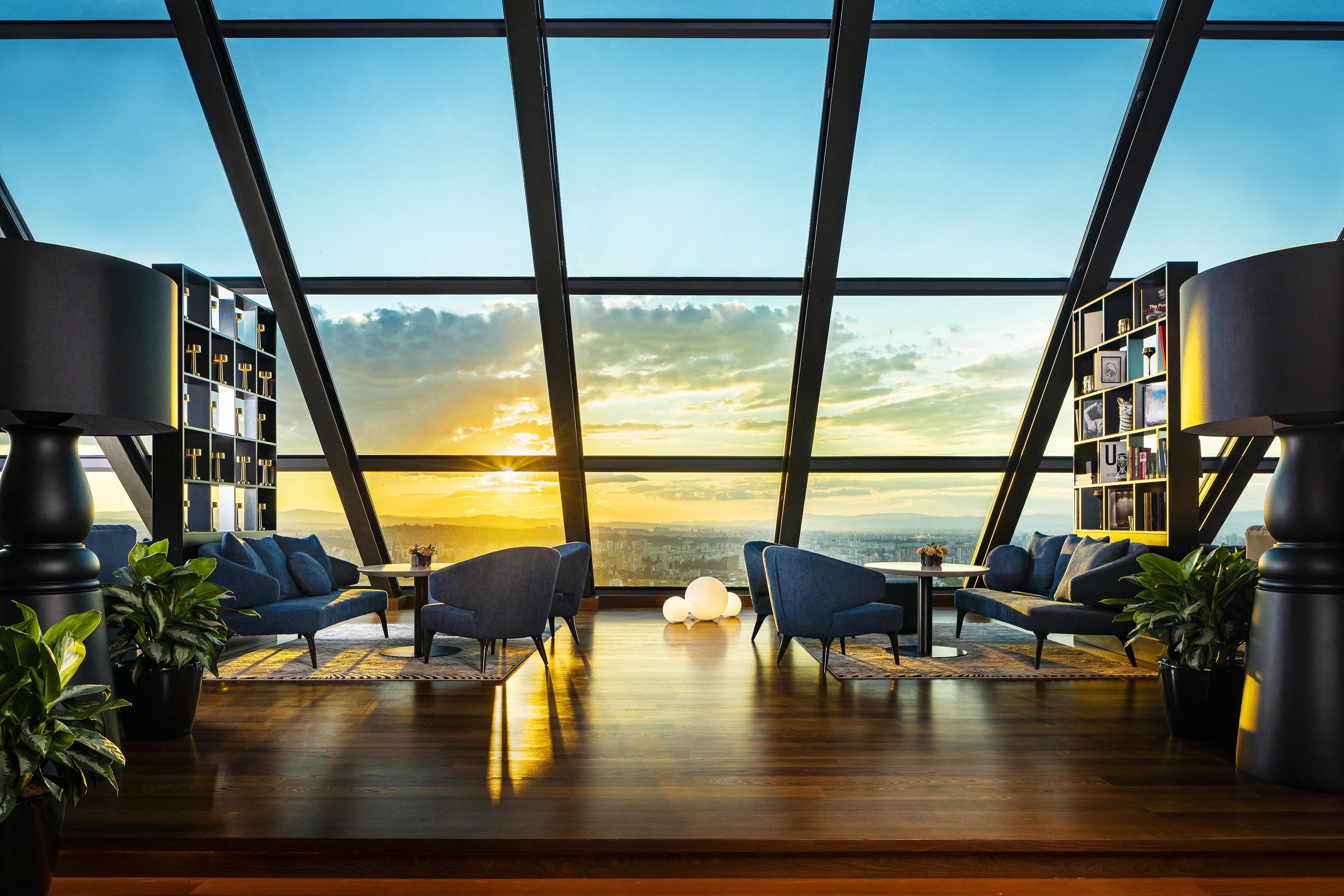 Grand Hotel Millennium 索菲亞 外观 照片 The photo depicts a modern, stylish interior of a high-rise space with a panoramic view through large glass windows. The scene features a cozy seating area with several blue armchairs arranged around a low table, creating a comfortable atmosphere. Th