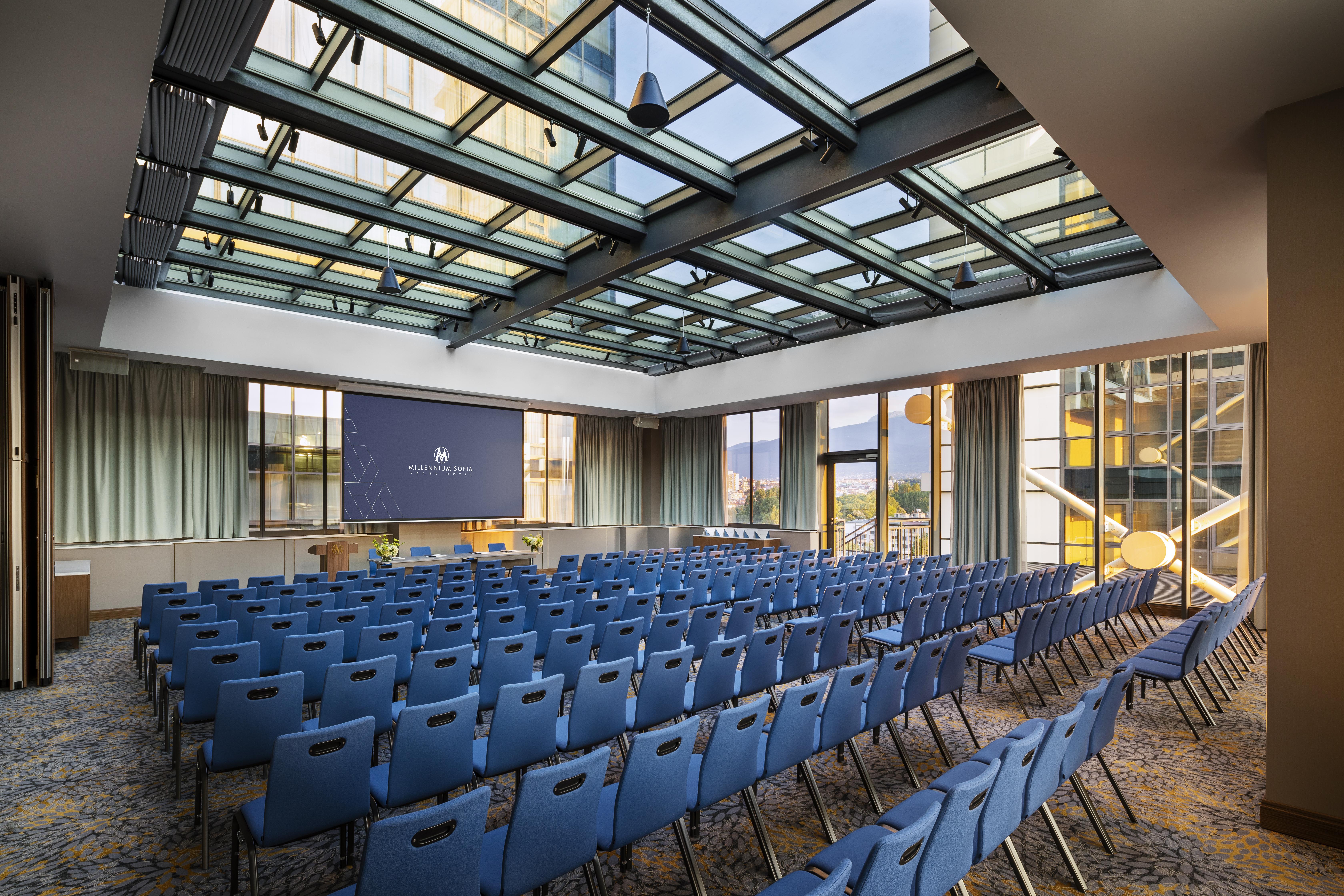 Grand Hotel Millennium 索菲亞 外观 照片 The photo shows a large conference room or meeting space. It features rows of blue chairs arranged in a theater style, facing a large screen at the front of the room. The room is well-lit, with a glass ceiling allowing natural light to filter in. The