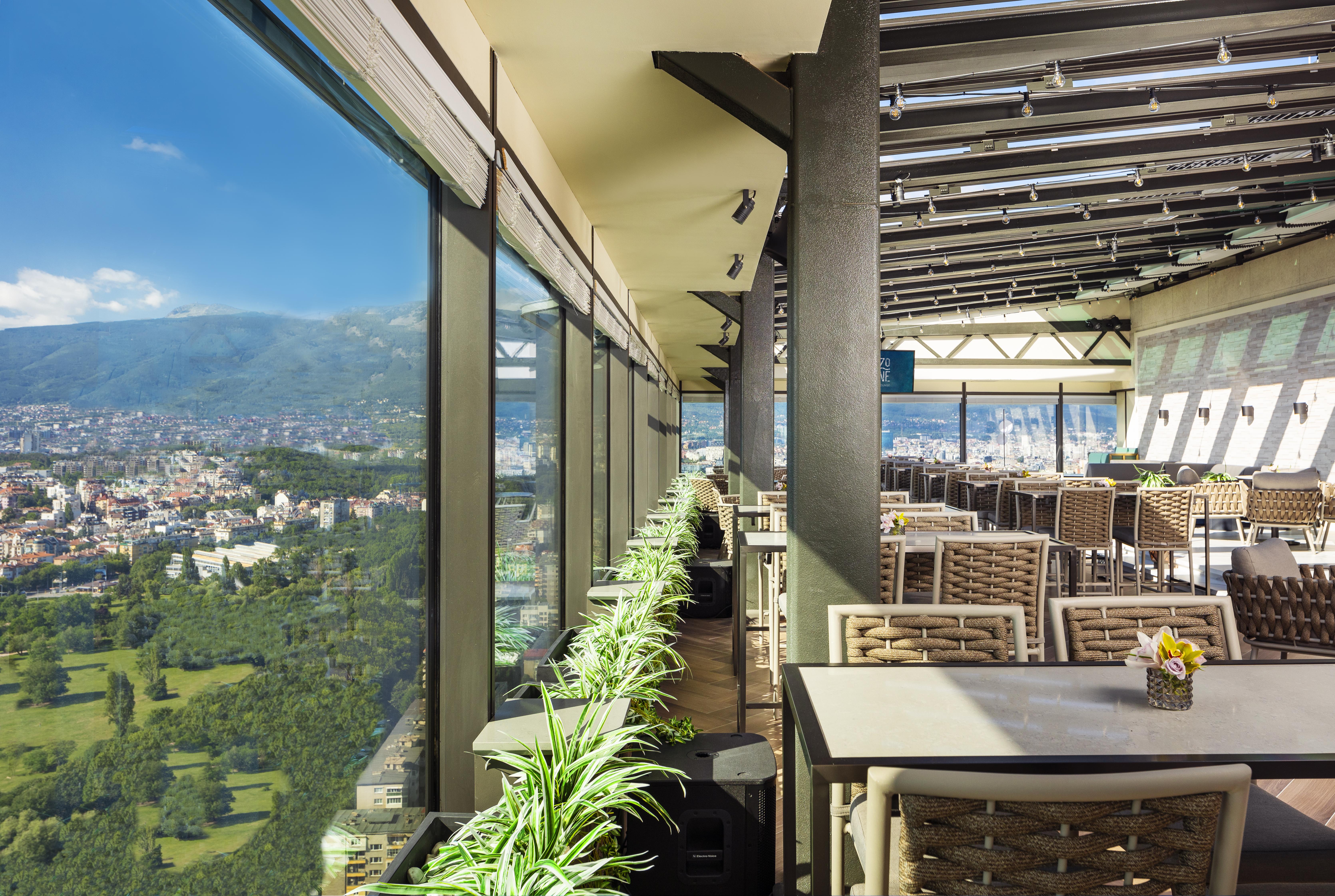 Grand Hotel Millennium 索菲亞 外观 照片 The photo shows a modern, spacious restaurant with large glass windows offering a panoramic view of a city and surrounding landscape. The interior features contemporary furniture, including stylish tables and chairs. There are potted plants along the